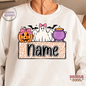 May include: White sweatshirt with a Halloween-themed design featuring a ghost, a pumpkin, and a cauldron. The design is on a leopard print background with the word "name" in black text.