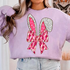 May include: A lavender sweatshirt featuring a pink bunny ear design. The bunny ears have black polka dots and pink paw prints. A pink and gold bow is tied at the base of the ears.