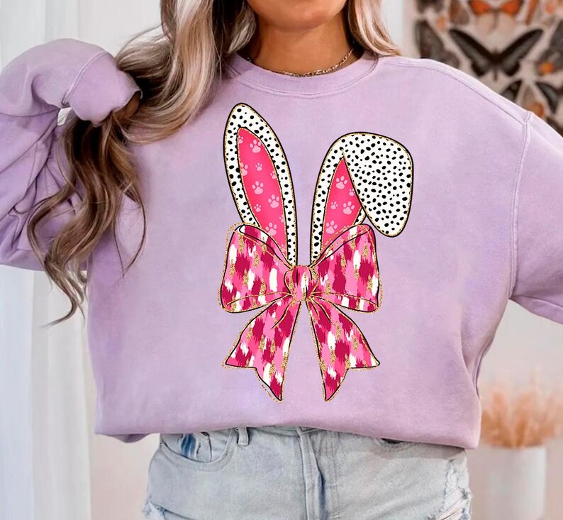 May include: A lavender sweatshirt featuring a pink bunny ear design. The bunny ears have black polka dots and pink paw prints. A pink and gold bow is tied at the base of the ears.