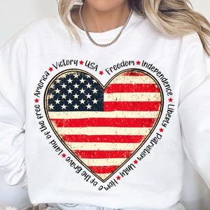 May include: White sweatshirt with a red, white, and blue heart-shaped American flag design. The heart is surrounded by the words "Home of the Brave, Land of the Free, America, Victory, USA, Freedom, Independence, Liberty, Patriotism, Unity".