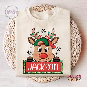 May include: A beige Christmas sweatshirt with a cartoon reindeer wearing a green and red hat. The reindeer has a red nose and the text "JACKSON" is printed on a green and white striped banner.