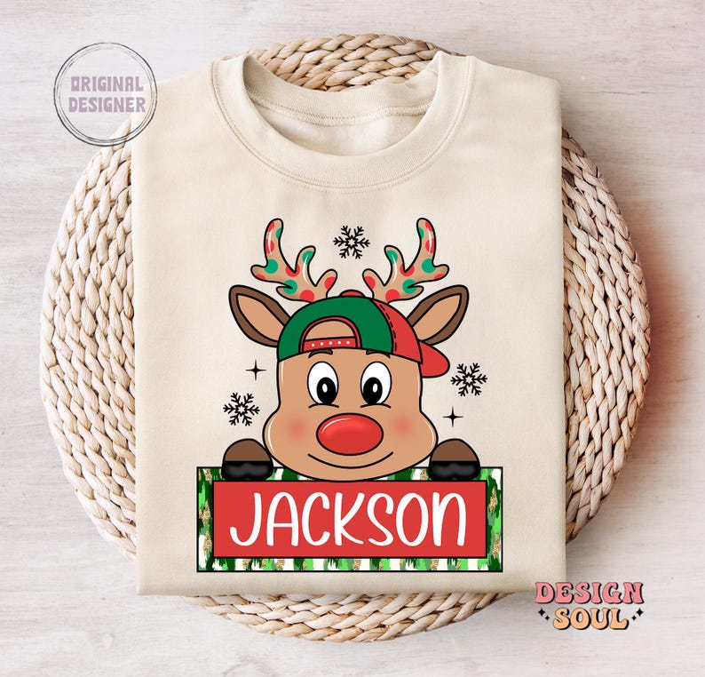 May include: A beige Christmas sweatshirt with a cartoon reindeer wearing a green and red hat. The reindeer has a red nose and the text "JACKSON" is printed on a green and white striped banner.