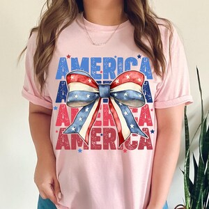 May include: Pink t-shirt with the word "AMERICA" in blue and red block letters. A patriotic bow graphic in red, white, and blue is centered on the shirt. The bow has a star pattern and is a festive design.