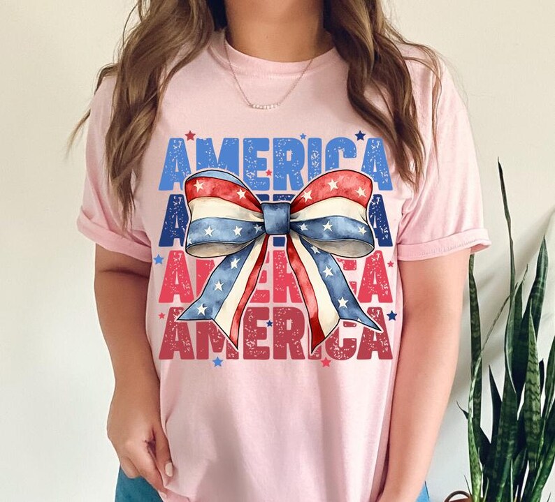 May include: Pink t-shirt with the word "AMERICA" in blue and red block letters. A patriotic bow graphic in red, white, and blue is centered on the shirt. The bow has a star pattern and is a festive design.