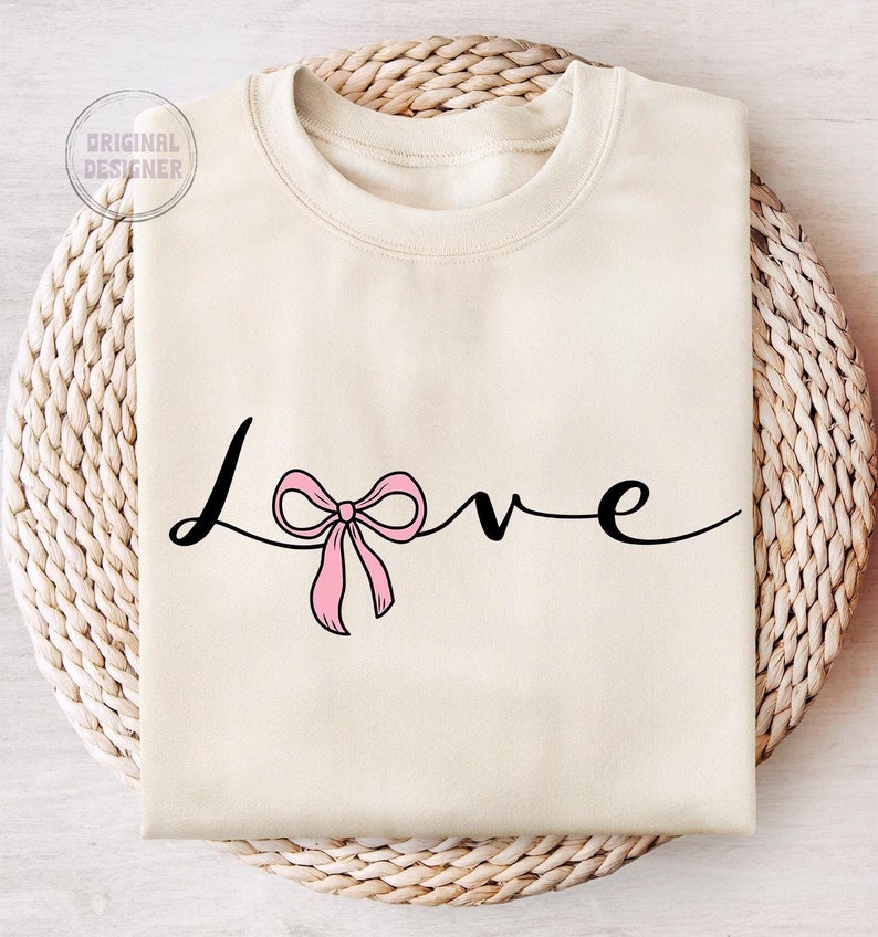 May include: A cream colored sweatshirt with the word "love" written in black cursive script. A pink bow is tied in the middle of the word "love".