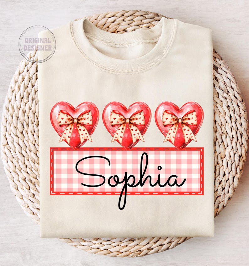 May include: A white sweatshirt with three red hearts with bows and a pink and white gingham banner with the name "Sophia" printed on it.