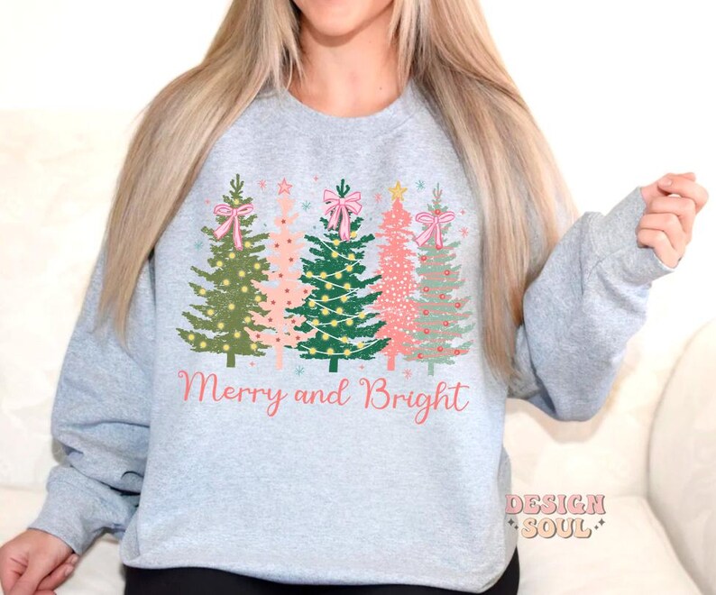 May include: A gray sweatshirt with a Christmas tree design and the text "Merry and Bright". The trees are decorated with pink bows, lights, and ornaments.