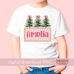 以下が含まれることがあります： ピンクと白のギンガムチェック柄の白いTシャツ。ピンクのリボンが付いた緑のクリスマスツリーが3本と、ピンクの文字で「アメリア」と書かれています。