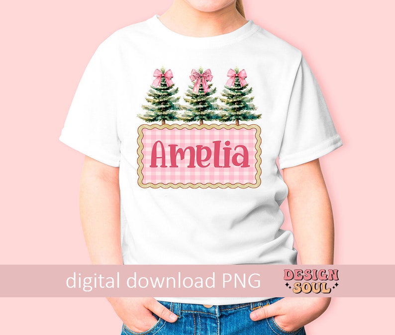 以下が含まれることがあります： ピンクと白のギンガムチェック柄の白いTシャツ。ピンクのリボンが付いた緑のクリスマスツリーが3本と、ピンクの文字で「アメリア」と書かれています。