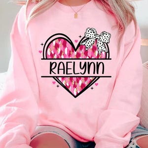 May include: Pink sweatshirt with a black heart design featuring a pink and gold leopard print. The heart has a black bow with white polka dots and the name "Raelynn" written across the center.