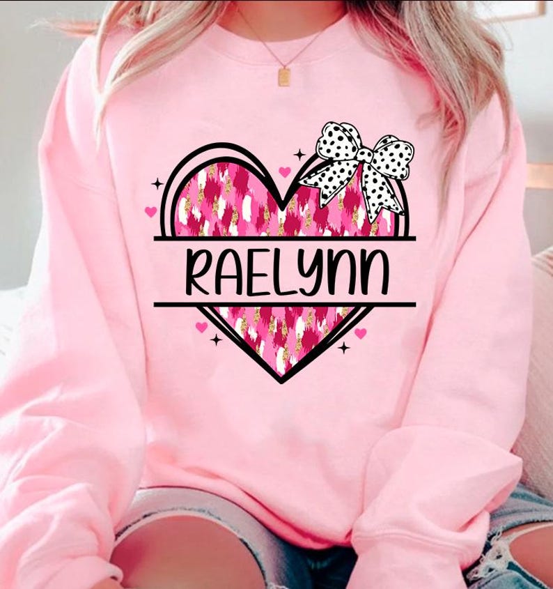 May include: Pink sweatshirt with a black heart design featuring a pink and gold leopard print. The heart has a black bow with white polka dots and the name "Raelynn" written across the center.