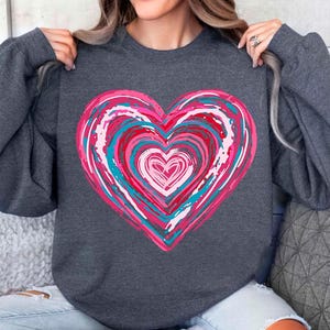 Pu&ograve; includere: Una felpa grigia scura con un grande disegno a forma di cuore rosa, blu e bianco. Il cuore &egrave; composto da cerchi concentrici di vernice.