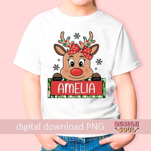 May include: White t-shirt with a cartoon reindeer wearing a red bow and holding a sign that says "Amelia". The reindeer has red antlers and a red nose. The sign is green and white with snowflakes and polka dots.