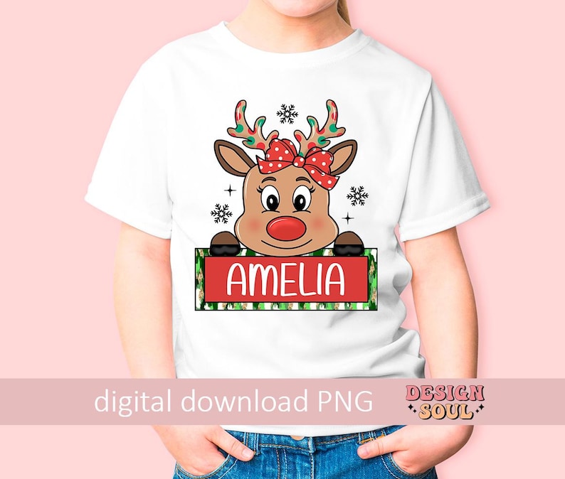 May include: White t-shirt with a cartoon reindeer wearing a red bow and holding a sign that says "Amelia". The reindeer has red antlers and a red nose. The sign is green and white with snowflakes and polka dots.