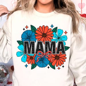 Pu&ograve; includere: Felpa bianca con un design floreale e la parola "MAMA" in lettere glitter nere. I fiori sono rossi, blu e verdi.