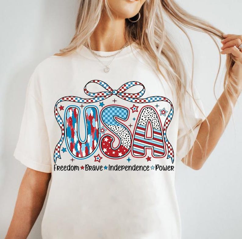 Peut inclure: T-shirt blanc avec un graphique USA rouge, blanc et bleu. Le graphique pr&eacute;sente un n&oelig;ud et des &eacute;toiles. Le texte sous le graphique indique "Freedom Brave Independence Power".