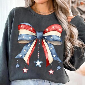 May include: A charcoal gray sweatshirt with a large red, white, and blue bow with stars printed on it. The bow has a blue center and is accented with dangling stars.