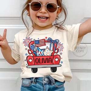 May include: A cream-colored t-shirt featuring a red truck graphic with an American flag, stars, and fireworks. The name "OLIVIA" is printed on the truck. The shirt is worn by a young child wearing sunglasses and blue jeans.
