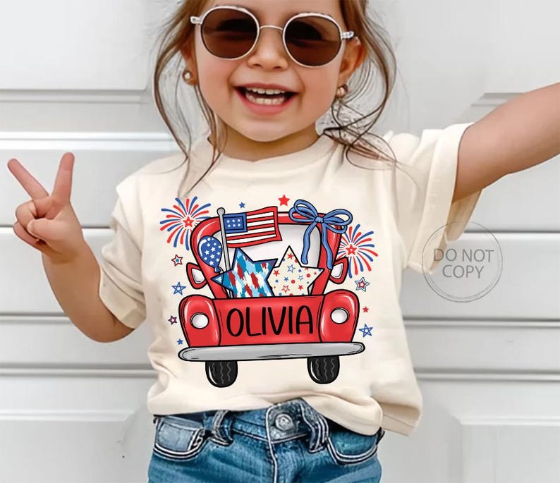 May include: A cream-colored t-shirt featuring a red truck graphic with an American flag, stars, and fireworks. The name "OLIVIA" is printed on the truck. The shirt is worn by a young child wearing sunglasses and blue jeans.