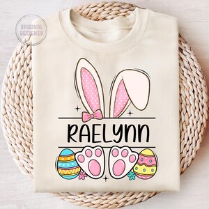 May include: A beige sweatshirt with a pink bunny ears design, the name "Raelynn" in black text, and two colorful Easter eggs.