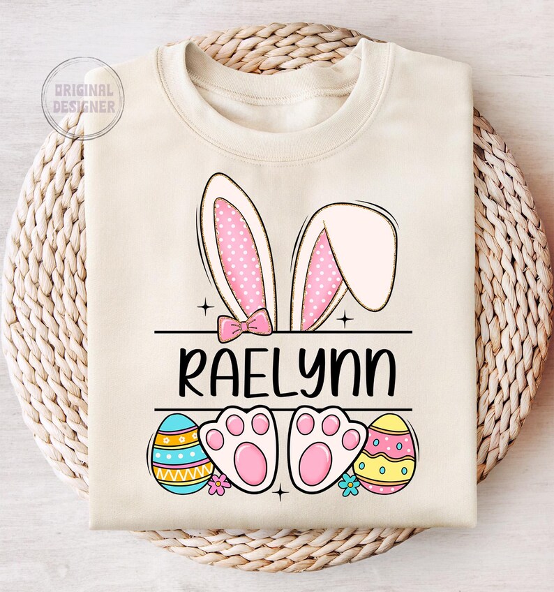 May include: A beige sweatshirt with a pink bunny ears design, the name "Raelynn" in black text, and two colorful Easter eggs.