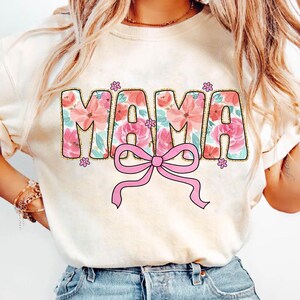 May include: A white t-shirt with a floral design that spells out "MAMA" in pink and gold glitter. A pink bow is centered below the word.