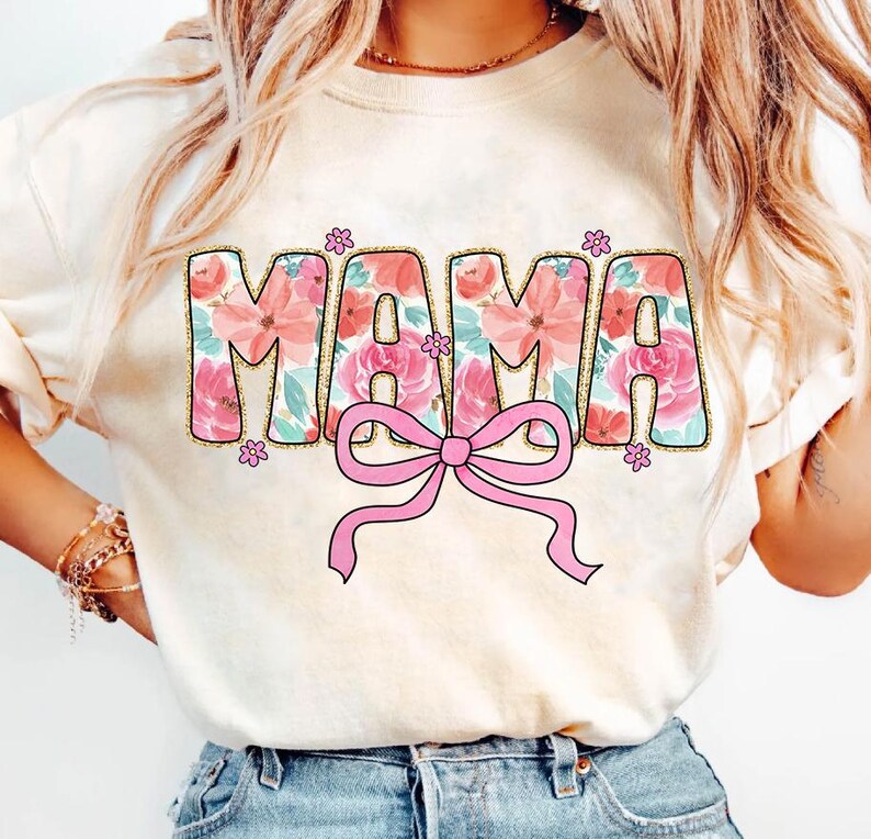May include: A white t-shirt with a floral design that spells out "MAMA" in pink and gold glitter. A pink bow is centered below the word.