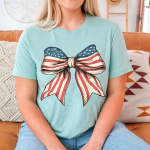 Peut inclure: Un t-shirt bleu clair avec un grand n&oelig;ud rouge, blanc et bleu imprim&eacute; sur le devant. Le n&oelig;ud est con&ccedil;u pour ressembler au drapeau am&eacute;ricain.