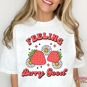 May include: White t-shirt with a retro-style graphic design featuring two red strawberries with green leaves and white daisies. The text "Feeling Berry Good" is written in a bold, retro font.