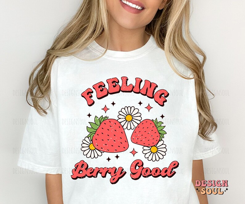 May include: White t-shirt with a retro-style graphic design featuring two red strawberries with green leaves and white daisies. The text "Feeling Berry Good" is written in a bold, retro font.
