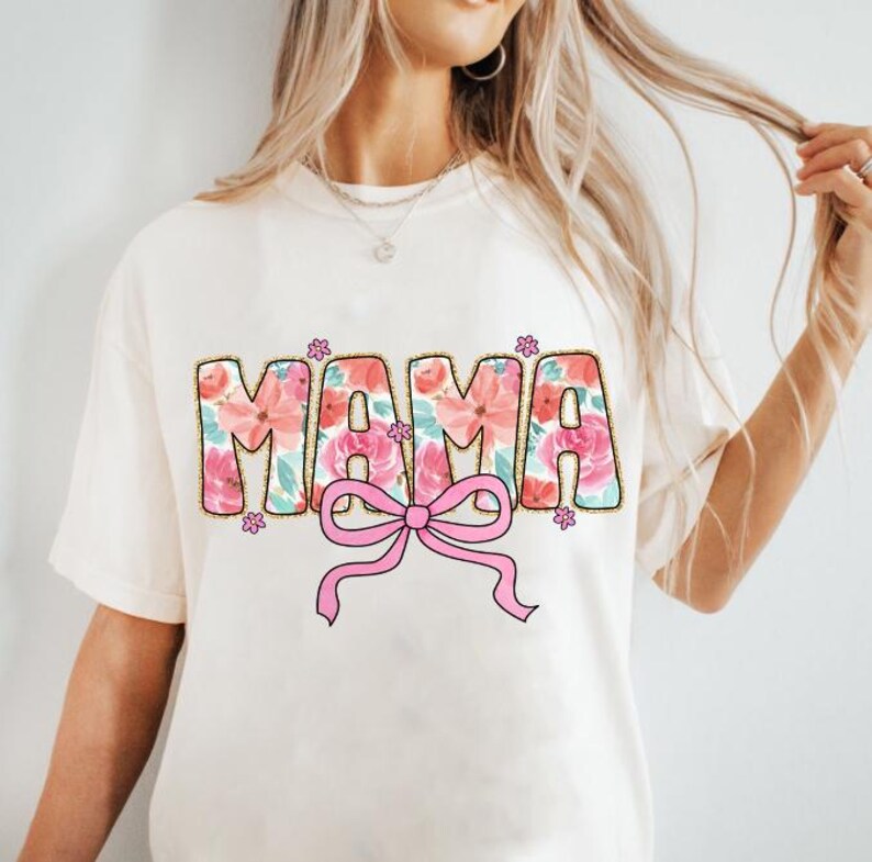 May include: White t-shirt with a floral print design that says "MAMA" with a pink bow underneath.