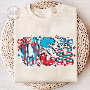 May include: A cream-colored sweatshirt with the word "USA" printed in large, colorful letters. The letters are decorated with red, white, and blue stars, stripes, and checkered patterns.  The letters are accented with red and blue bows.