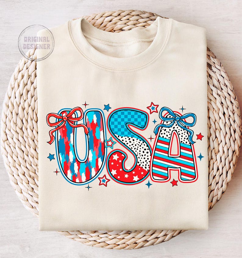 May include: A cream-colored sweatshirt with the word "USA" printed in large, colorful letters. The letters are decorated with red, white, and blue stars, stripes, and checkered patterns.  The letters are accented with red and blue bows.