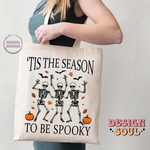 May include: A white tote bag with the text "'Tis the season to be spooky" printed on it. The design features three skeletons dancing with pumpkins and autumn leaves. The text "Design Soul" is printed in pink on the bottom right corner of the bag.