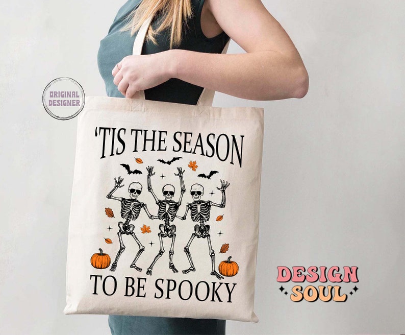 May include: A white tote bag with the text "'Tis the season to be spooky" printed on it. The design features three skeletons dancing with pumpkins and autumn leaves. The text "Design Soul" is printed in pink on the bottom right corner of the bag.