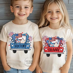 May include: Two children wearing cream-colored t-shirts with a patriotic design. The shirts feature a cartoon truck carrying stars, fireworks, and an American flag. One shirt says "AXTON" and the other says "OLIVIA".