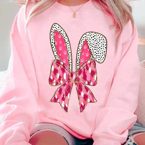 May include: Pink sweatshirt featuring a pink and gold bunny ears and bow design. The bow has a pink and white leopard print pattern.