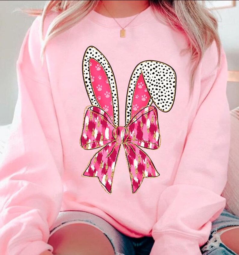 May include: Pink sweatshirt featuring a pink and gold bunny ears and bow design. The bow has a pink and white leopard print pattern.