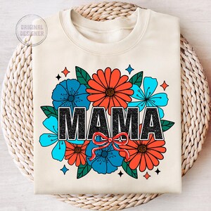 Pu&ograve; includere: Una felpa color crema con un design floreale e la parola "MAMA" in lettere nere. I fiori sono rossi, blu e verdi.