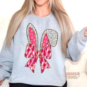 May include: A grey sweatshirt featuring a pink and gold bunny ear design. The ears have a black and white polka dot pattern and pink paw prints. The bunny ears are tied with a pink and gold bow.