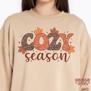 May include: A beige sweatshirt with the words "COZY season" in a brown and orange glittery font, decorated with autumn leaves. The sweatshirt has a crew neck and long sleeves, perfect for fall.