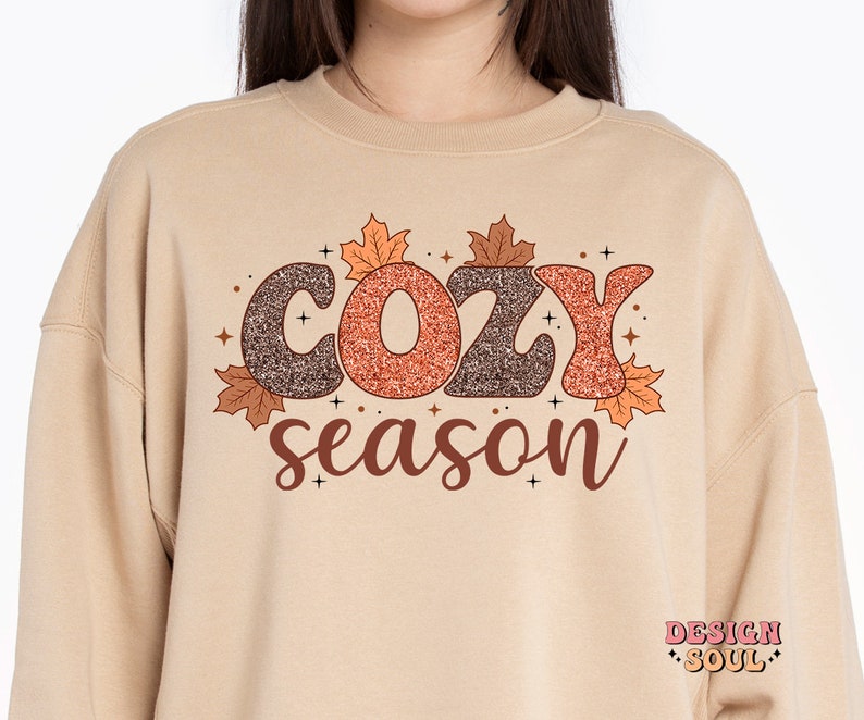 May include: A beige sweatshirt with the words "COZY season" in a brown and orange glittery font, decorated with autumn leaves. The sweatshirt has a crew neck and long sleeves, perfect for fall.