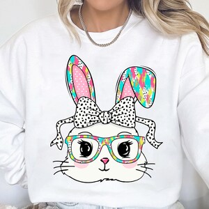 May include: White sweatshirt with a colorful cartoon bunny wearing glasses and a bow. The bunny's ears are pink and white with black polka dots. The bow is black with white polka dots. The glasses are rainbow colored.