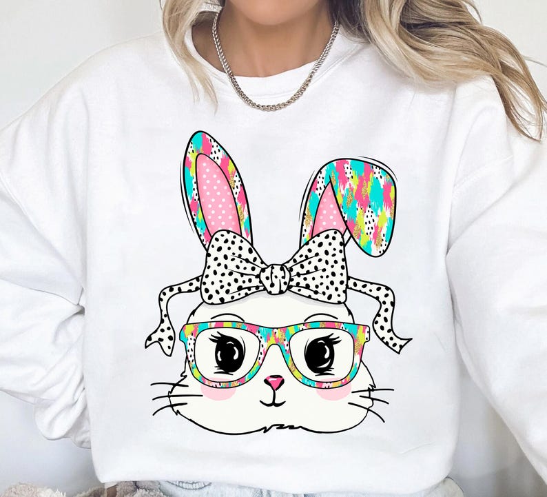 May include: White sweatshirt with a colorful cartoon bunny wearing glasses and a bow. The bunny's ears are pink and white with black polka dots. The bow is black with white polka dots. The glasses are rainbow colored.