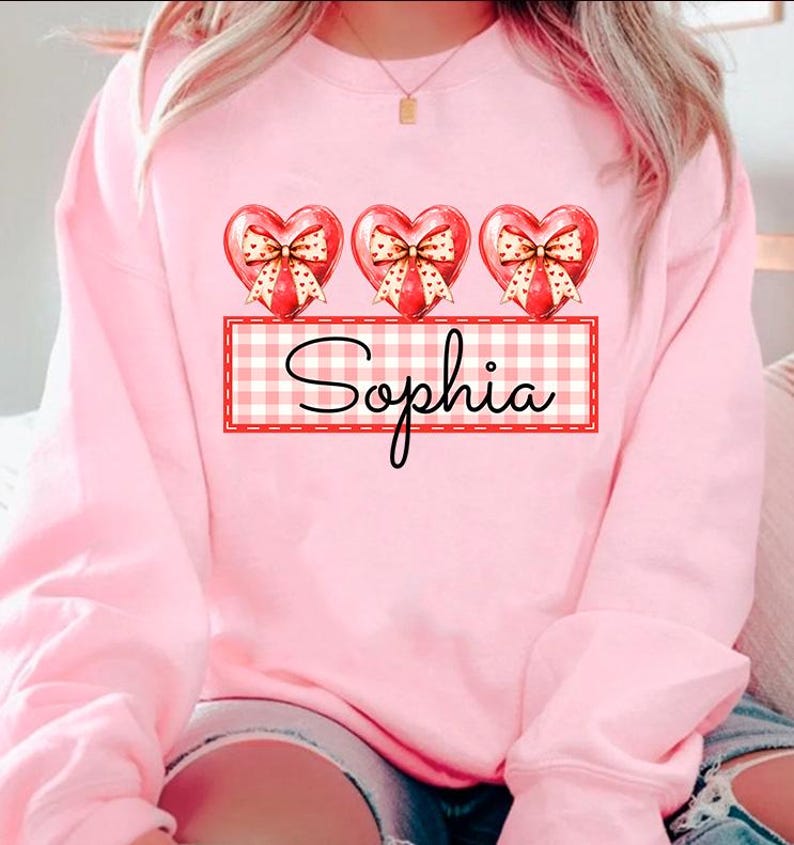 May include: Pink sweatshirt with three red hearts with bows and a red and white checkered banner with the name "Sophia" printed on it.