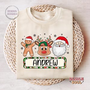 May include: A beige Christmas sweater with a reindeer, gingerbread man, and Santa Claus design. The sweater has a green, red, and white striped banner with the name "ANDREW" printed on it.