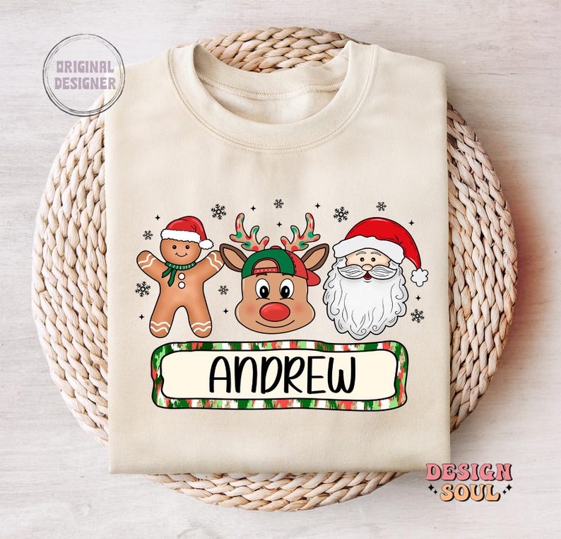 May include: A beige Christmas sweater with a reindeer, gingerbread man, and Santa Claus design. The sweater has a green, red, and white striped banner with the name "ANDREW" printed on it.
