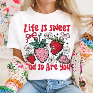 May include: White t-shirt with a red graphic that says "Life is sweet and so are you". The graphic also features strawberries, flowers, and bows.