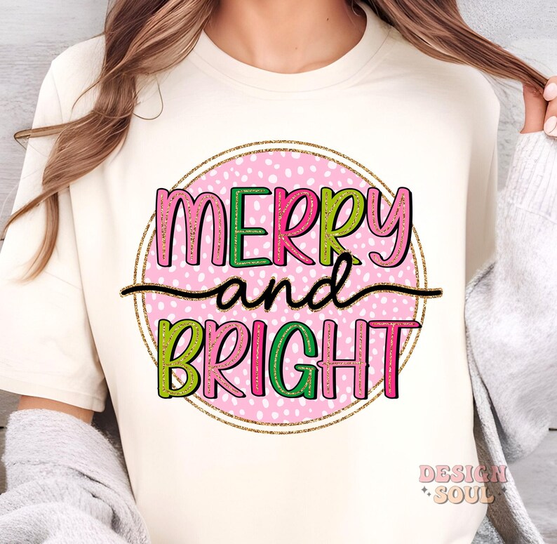May include: Cream-colored t-shirt with a festive graphic. The design features the words "MERRY and BRIGHT" in pink, green, and black, set within a pink polka-dotted circle with a gold glitter outline. The shirt has a crew neck.