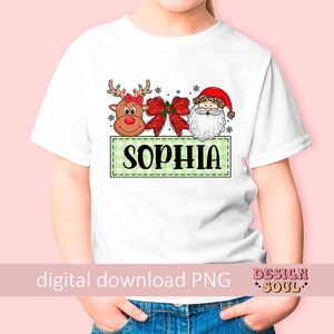 May include: White t-shirt with a green and white checkered box with the name "Sophia" in red letters. The box contains a cartoon reindeer with a red bow, a red bow, and a cartoon Santa Claus with a white beard.